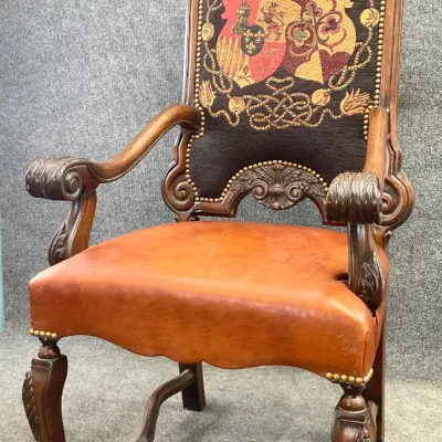 Sale Photo Thumbnail #96: Ornate Spanish Baroque-style armchair featuring a carved wood frame, a brown leather seat with brass nailhead trim, and an embroidered tapestry backrest depicting a royal coat of arms.