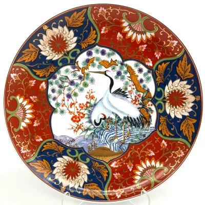 Sale Photo Thumbnail #58: Japanese Imari porcelain charger featuring a central motif of two cranes amidst pine and plum blossoms, surrounded by alternating red and blue floral panels with gold gilt accents.