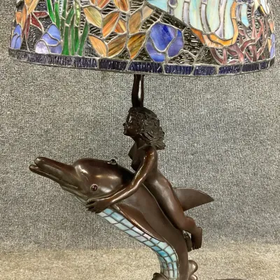 Sale Photo Thumbnail #24: Tiffany-style table lamp featuring a bronze-finished base of a mermaid riding a dolphin, topped with a colorful stained glass shade depicting an underwater scene with fish and aquatic plants.