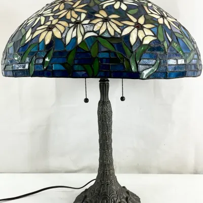 Sale Photo Thumbnail #10: Tiffany-style stained glass table lamp featuring a floral daisy motif in shades of blue, green, and white, mounted on an ornate bronze-finished base with dual pull chains.