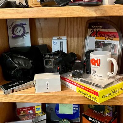 Wooden shelving unit filled with vintage electronics, cameras, blank media, and novelty items, including an Olympus camera, a Tiffen...