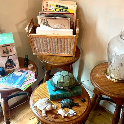 Assorted home decor items displayed on wooden stools, including a stained glass turtle lamp, a wicker magazine rack filled with books,...