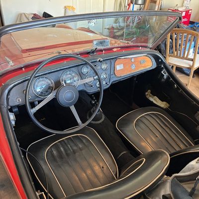 Sale Photo Thumbnail #3: Interior view of a vintage red convertible sports car, featuring black vinyl bucket seats with white piping, a classic banjo-style steering wheel, and a detailed dashboard.