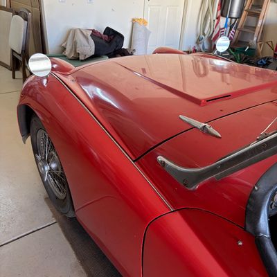 Sale Photo Thumbnail #11: Vintage red Triumph TR3 roadster, showing the front fender, wire wheel, hood, and fender-mounted side mirror.