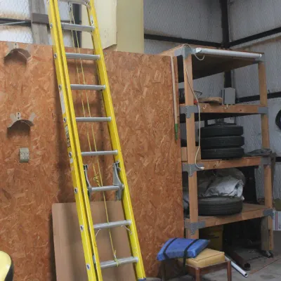 Yellow fiberglass extension ladder leaning against a wooden wall in a workshop, alongside a wooden shelving unit holding tires.