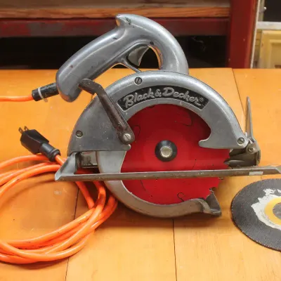 Vintage Black & Decker circular saw with a cast metal body, orange power cord, and an extra abrasive cutting disc.
