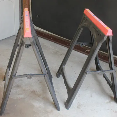 Pair of folding plastic sawhorses with black frames and orange top rails, showing light wear from use.