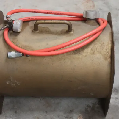 Vintage portable air tank with a brass-colored finish, featuring an attached red hose, pressure gauge, and carrying handle.