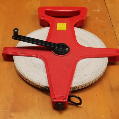 Cen-Tech open reel fiberglass measuring tape with a red plastic frame and black hand crank, resting on a wooden surface.