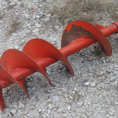 Vintage orange metal auger attachment, showing some wear and paint chipping, resting on a gravel surface.