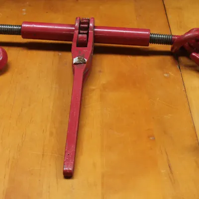 Red painted steel ratchet chain binder with grab hooks, showing minor wear from use.