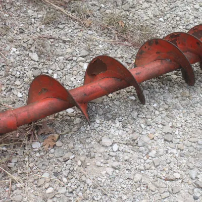 Vintage red metal auger bit showing wear and chipped paint, resting on a gravel surface.