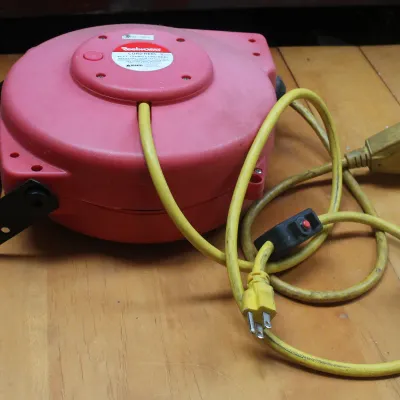 Reelworks retractable extension cord reel with a red plastic housing, black mounting bracket, and a yellow 12 AWG cord.