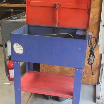 Vintage electric-powered 10-gallon parts washer with a blue body, red lid, and lower storage shelf.