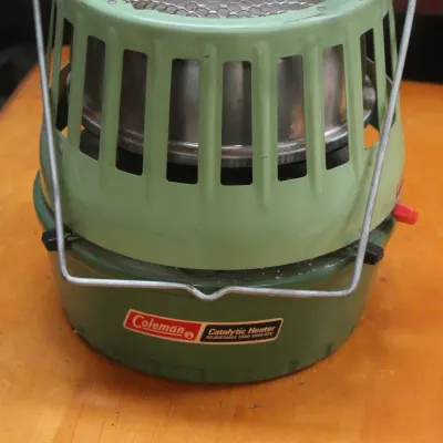 Vintage Coleman adjustable catalytic heater in two-tone green with a wire carrying handle, showing some light wear from use.