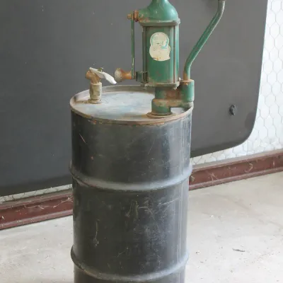 Vintage Quaker State green hand pump mounted on a black metal oil drum, showing age-appropriate wear and patina.