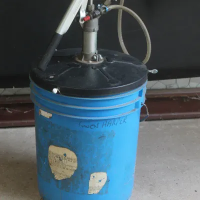 Manual bucket pump mounted on a blue plastic five-gallon bucket, showing signs of use with worn labels.