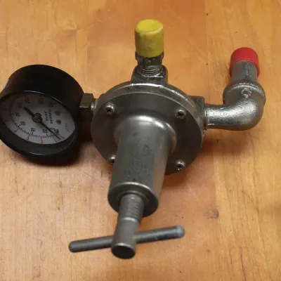 Vintage metal pressure regulator with attached pressure gauge, T-handle adjustment screw, and protective plastic caps on the ports.