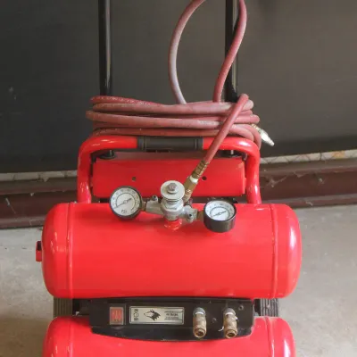 Rockworth Classic Air portable twin-tank air compressor in red, featuring a black handle, dual pressure gauges, and an attached red hose.