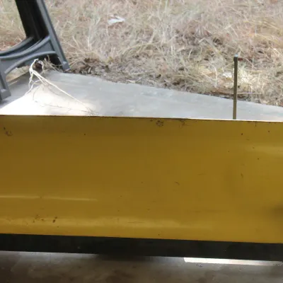 Yellow John Deere garden tractor snow plow attachment, showing some wear and light rust from use.