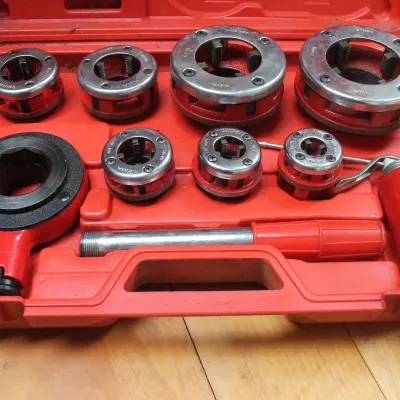 Manual pipe threading tool set with multiple die heads, ratchet handle, and accessories, housed in a red molded plastic carrying case.