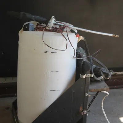 Portable chemical or spot sprayer system featuring a white plastic tank with volume markings, mounted on a black metal frame with an...