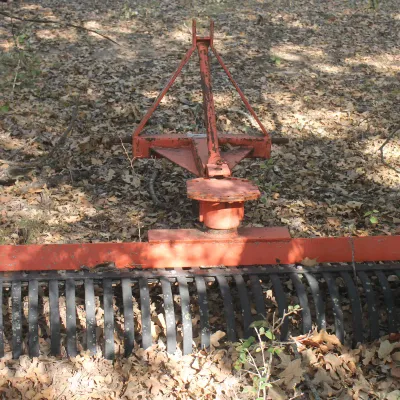 Three-point hitch landscape rake with an orange frame and curved black tines, showing some wear and weathering from outdoor storage.