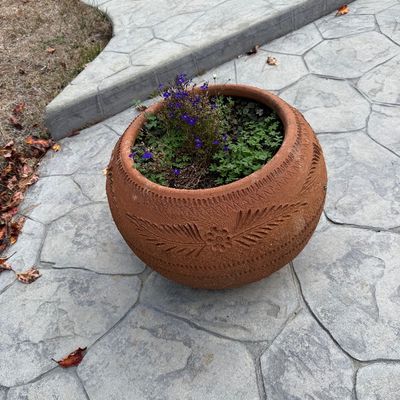 Large round terracotta planter featuring carved floral and leaf motifs, planted with small purple flowers and greenery.