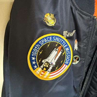 Vintage navy blue flight jacket featuring a large 100th Space Shuttle Mission embroidered patch and several space-themed pins.