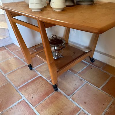 Mid-century modern wooden drop-leaf rolling bar cart or tea cart, featuring a lower shelf and angled legs on casters, styled with a...