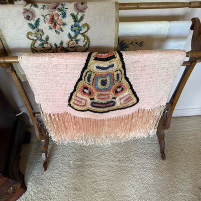 Vintage wooden quilt rack displaying a collection of embroidered and needlepoint textiles, including a pink fringed piece with a central...