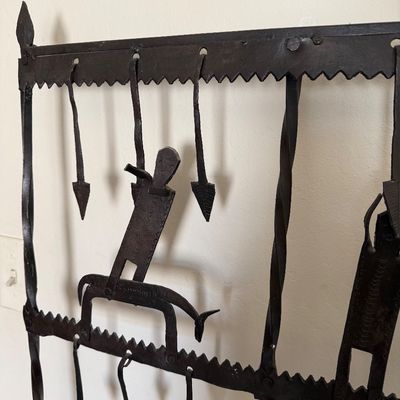 Hand-forged wrought iron wall art panel featuring stylized figures, arrows, and twisted metal bars in a rustic, folk art style.