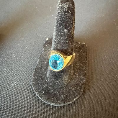 Gold ring featuring a round, faceted light blue gemstone, displayed on a black velvet ring stand.