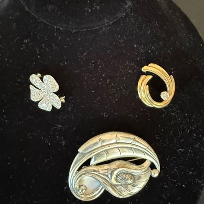 Three vintage brooches displayed on a black velvet stand, including a silver, four-leaf clover, a gold-tone swirl with a rhinestone, and...