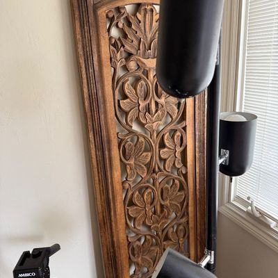 Mid-century modern three-light tension pole lamp in matte black, shown alongside an ornate carved wood wall panel and an Ambico camera...