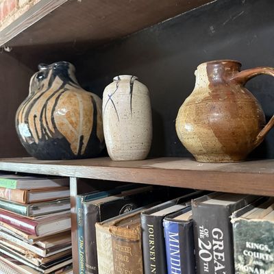 Three studio art pottery vessels, including a pitcher and two vases with earthy glazes, displayed on a wooden bookshelf above a...
