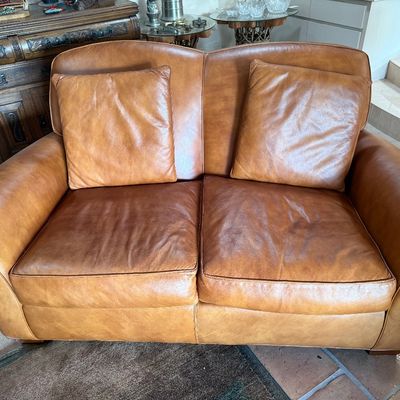 Tan leather loveseat with rolled arms and matching throw pillows, showing some natural wear and patina.