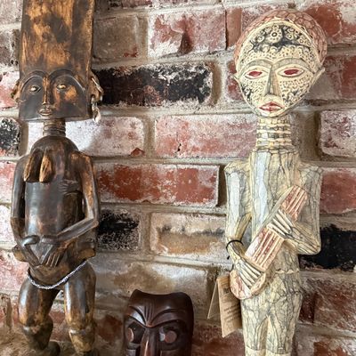 Two hand-carved African-style wooden statues and a small carved wooden mask displayed against a brick wall.