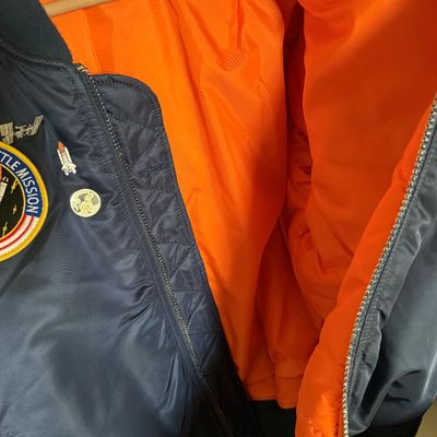 Alpha Industries MA-1 bomber jacket in navy blue with a bright orange lining, featuring space mission patches and pins.