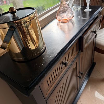Wooden bar cabinet with a black top, featuring slatted doors and drawers, styled with a vintage brass ice bucket and glassware.