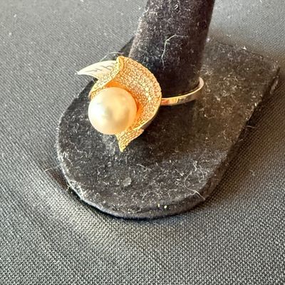 Gold ring featuring a single cultured pearl nestled in a textured, leaf-like setting, displayed on a black velvet ring stand.