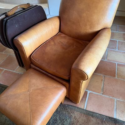 Tan leather armchair and matching ottoman with cross-stitch detailing, showing minor wear from use.