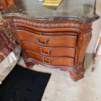 Traditional three-drawer nightstand featuring a dark marble top, ornate wood carvings, and curved drawer fronts with metal pull handles.