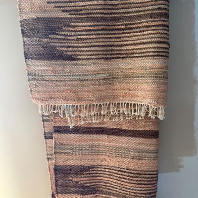 Woven rag rug with a fringed edge, featuring horizontal stripes in shades of pink, brown, and tan, hanging over a metal rod.