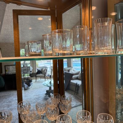 Assorted crystal glassware, including cut-glass wine goblets and ribbed tumblers, displayed on glass shelves inside a lighted curio cabinet.