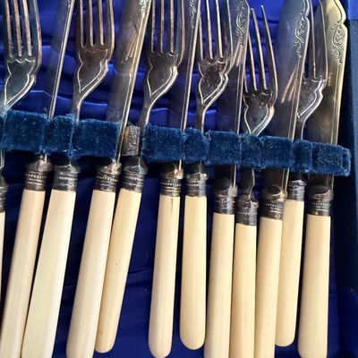 Antique silver-plated fish cutlery set with bone or celluloid handles, featuring ornate engraved details on the blades and tines,...