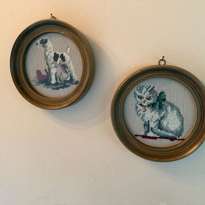 Pair of vintage needlepoint portraits featuring a dog and a cat, housed in round, gold-painted wooden frames with hanging loops.