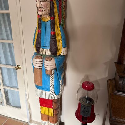 Hand-carved and painted wooden cigar store Indian statue, standing beside a vintage red gumball machine on a black pedestal stand.