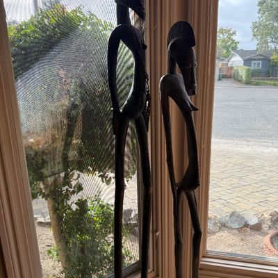 Pair of tall, slender African-style hand-carved wooden statues depicting stylized human figures, displayed near a window.