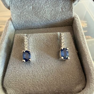 Pair of white gold or platinum drop earrings, each featuring an oval-cut blue sapphire suspended from a diamond-accented bale, presented...